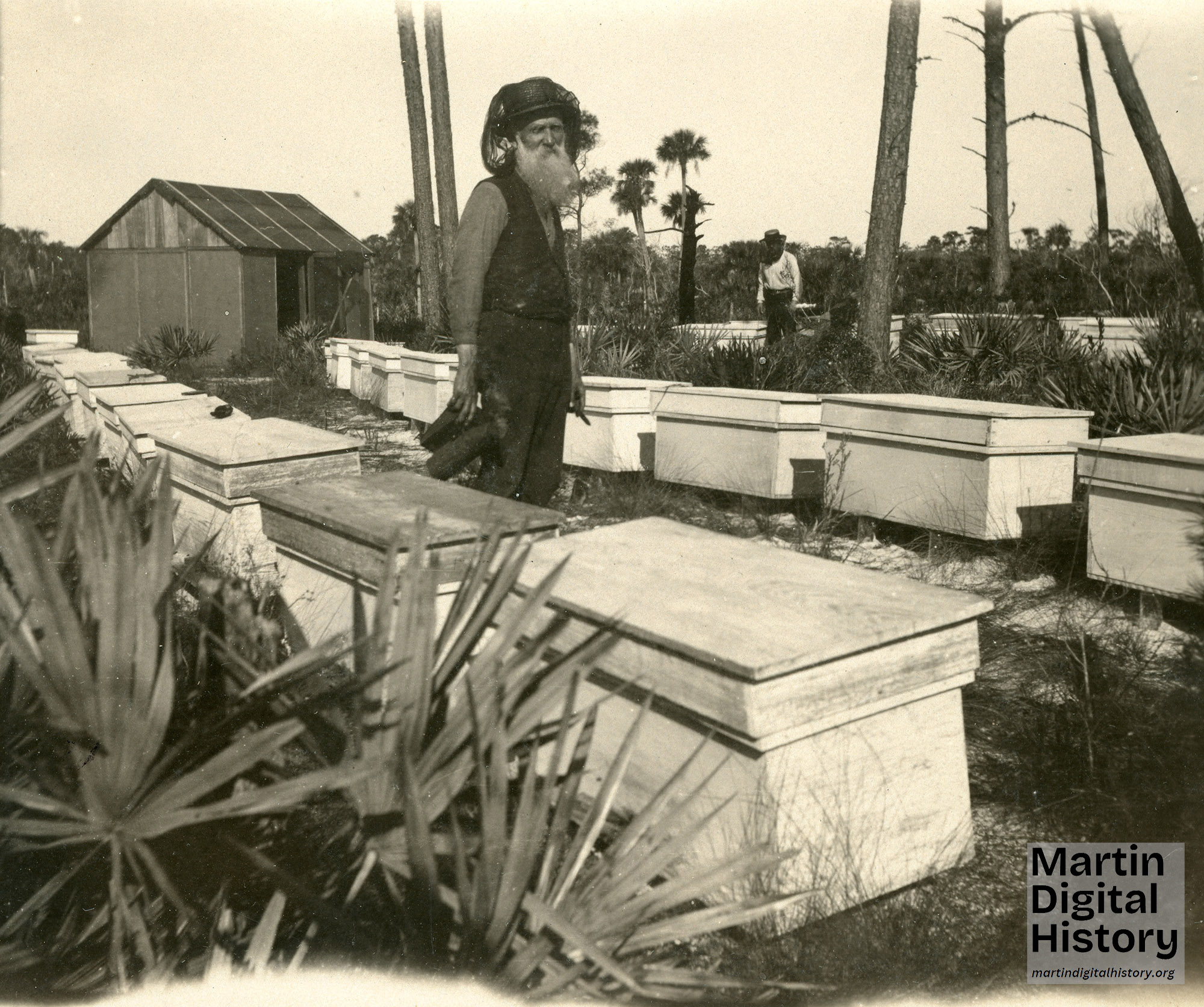 Colonel Oscar Ogden Poppleton stands between rows of beehive boxes holding a bee smoker.